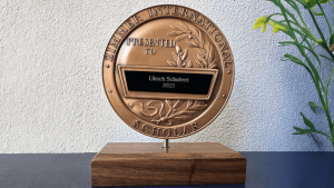 Bronzene Plakette des Zimmer International Scholar Award, überreicht an Ulrich Schubert im Jahr 2025, montiert auf einem Holzsockel.