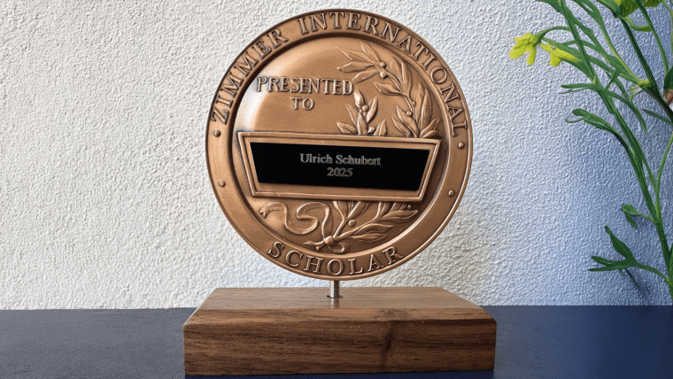 Bronzene Plakette des Zimmer International Scholar Award, überreicht an Ulrich Schubert im Jahr 2025, montiert auf einem Holzsockel.